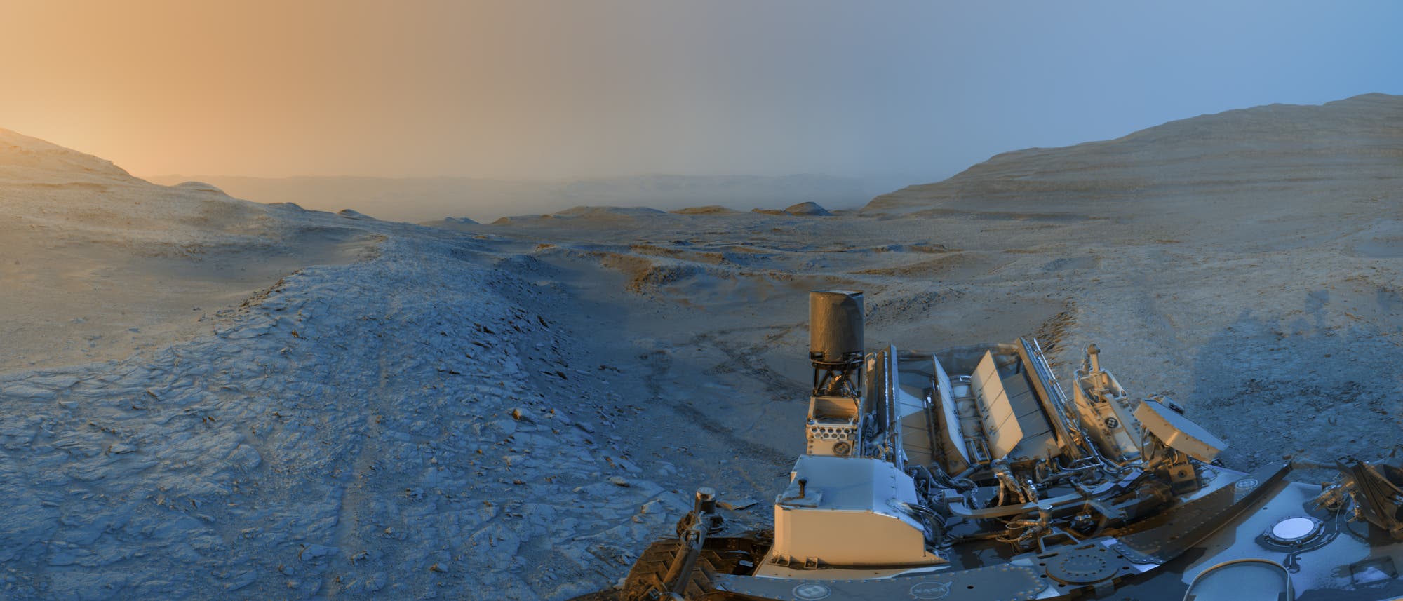 Eine Panoramaaufnahme der Marsoberfl&auml;che, aufgenommen von einem Rover. Im Vordergrund sind Teile des Rovers sichtbar, w&auml;hrend sich im Hintergrund eine weite, felsige Landschaft erstreckt. Der Himmel zeigt einen Farbverlauf von orange zu blau, was auf einen Sonnenaufgang oder Sonnenuntergang hindeutet. Die Szene vermittelt den Eindruck von Ruhe und Weite auf dem Mars.