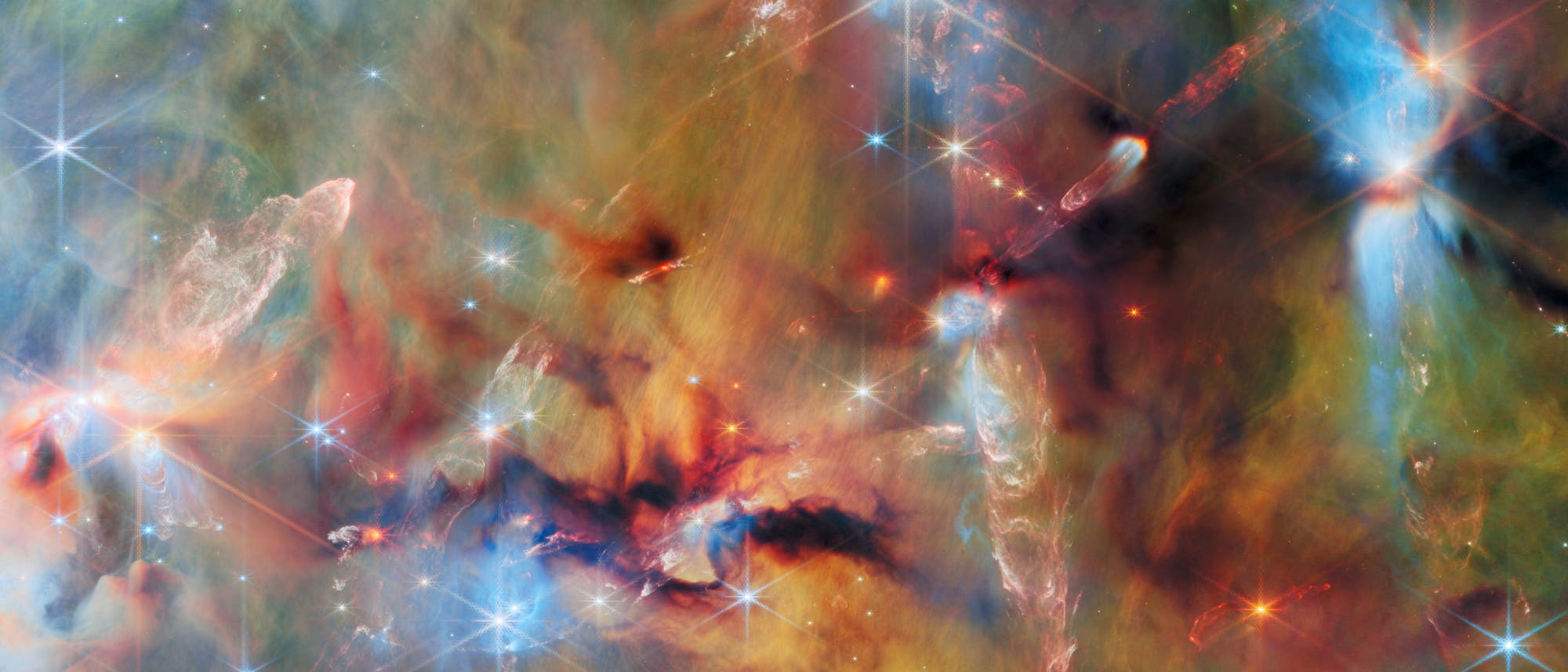 Ein farbenfrohes astronomisches Bild zeigt eine dichte Ansammlung von Sternen und leuchtenden Gaswolken im Weltraum. Verschiedene Farben wie Blau, Rot und Gelb mischen sich und bilden ein komplexes Muster aus Nebeln und Licht. Helle Sterne mit Lichtstrahlen sind &uuml;ber das Bild verteilt, w&auml;hrend die Gaswolken in unterschiedlichen Formen und Intensit&auml;ten leuchten. Das Bild vermittelt die Weite und Sch&ouml;nheit des Universums.