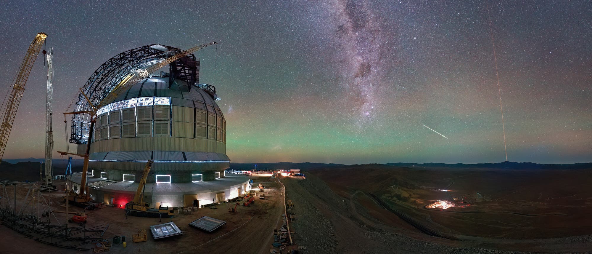 Ein großes Observatorium mit einer Kuppel und Baukränen steht in einer Wüstenlandschaft unter einem klaren Nachthimmel. Die Milchstraße ist deutlich sichtbar, und ein heller Meteor zieht eine Linie am Himmel. Im Hintergrund sind Berge und Lichter einer entfernten Siedlung zu sehen. Die Szene vermittelt eine ruhige, aber beeindruckende Atmosphäre der Astronomie und Technik.