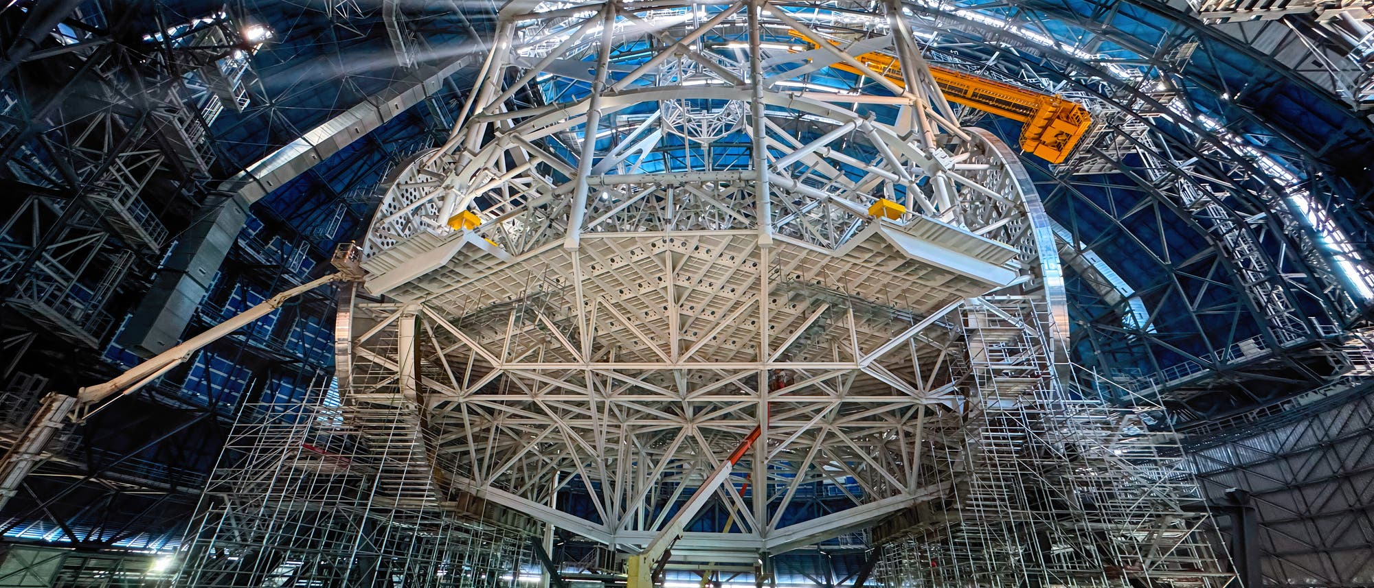 Innenansicht einer gro&szlig;en, industriellen Struktur mit einem komplexen Netzwerk aus Metalltr&auml;gern und Ger&uuml;sten. Sonnenstrahlen dringen durch die obere &Ouml;ffnung ein und beleuchten die Konstruktion. Ein orangefarbener Kran ist in der Mitte sichtbar, umgeben von zahlreichen Plattformen und Leitern. Die Szene vermittelt den Eindruck einer Baustelle oder eines technischen Projekts in einem gro&szlig;en Geb&auml;ude oder einer Kuppel.