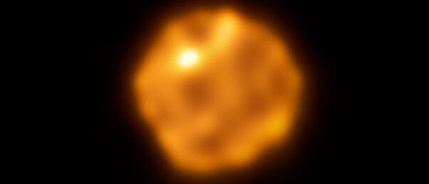 Ein verschwommenes astronomisches Bild zeigt einen orangefarbenen, leuchtenden Stern vor einem schwarzen Hintergrund. Der Stern hat eine unregelm&auml;&szlig;ige Form mit helleren und dunkleren Bereichen, die auf Oberfl&auml;chenstrukturen oder Temperaturunterschiede hinweisen k&ouml;nnten. Das Bild vermittelt den Eindruck von Hitze und Energie, typisch f&uuml;r stellare Objekte.