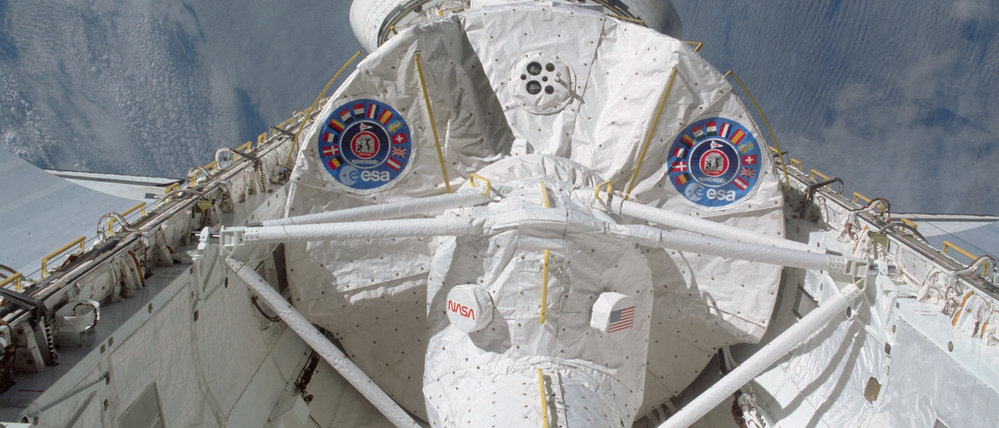 Ein Spaceshuttle im Weltraum, fotografiert von oben. Das Shuttle zeigt die Logos der NASA und ESA auf der Ladebucht. Im Hintergrund ist die Erde mit Wolkenformationen sichtbar. Die Struktur des Shuttles ist detailliert, mit sichtbaren technischen Komponenten und der US-amerikanischen Flagge. Ein Spaceshuttle im Weltraum, fotografiert von oben. Das Shuttle zeigt die Logos der NASA und ESA auf der Ladebucht. Im Hintergrund ist die Erde mit Wolkenformationen sichtbar. Die Struktur des Shuttles ist detailliert, mit sichtbaren technischen Komponenten und der US-amerikanischen Flagge.