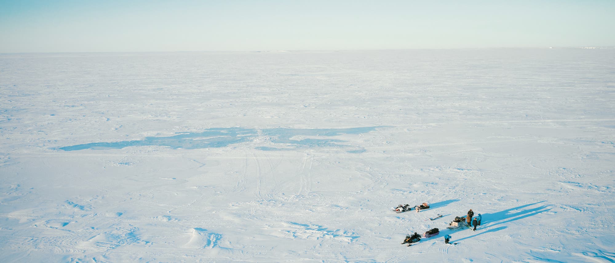 Eine weite, schneebedeckte Landschaft unter klarem, blauem Himmel. Im Vordergrund sind mehrere Schneemobile und Personen zu sehen, die auf der eisigen Fläche stehen. Die Szene vermittelt eine ruhige, abgelegene Atmosphäre in einer kalten, arktischen Umgebung.
