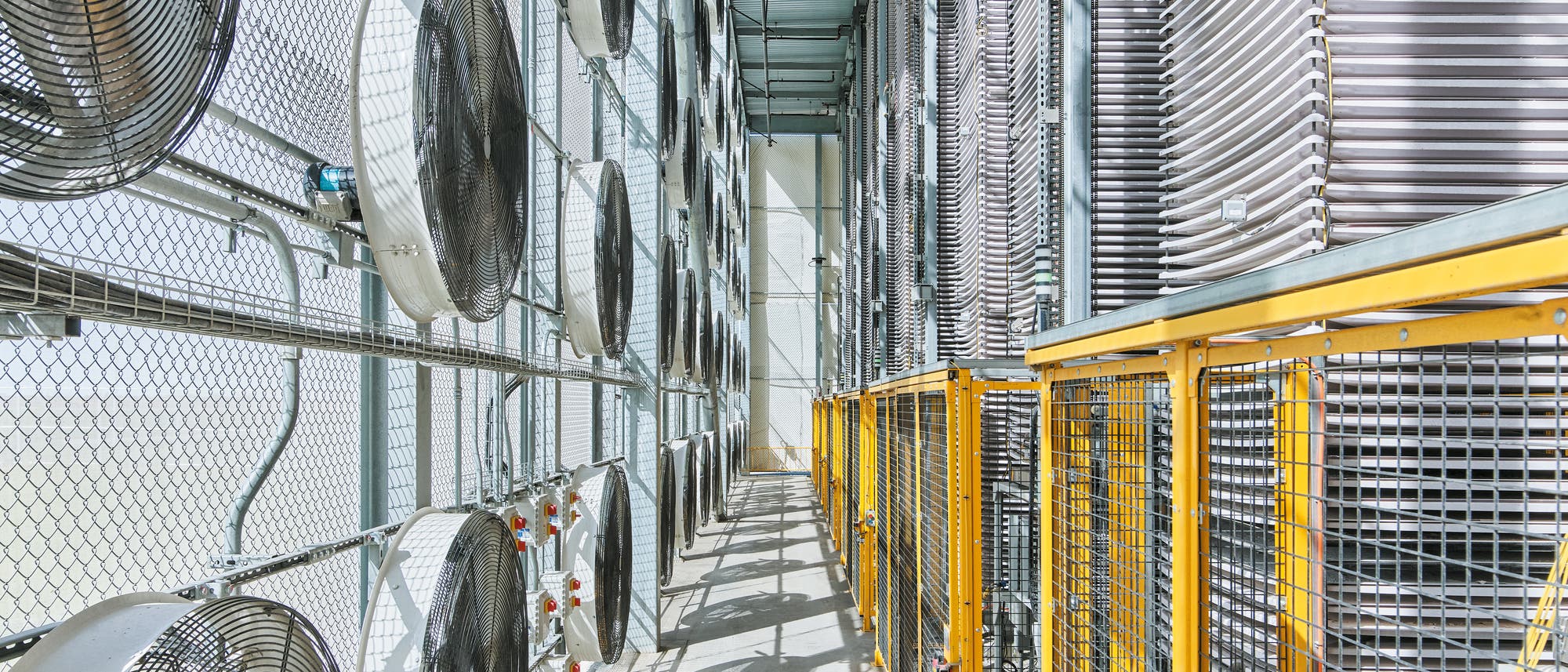 Foto aus dem Innern einer Anlage zum Entzug von Kohlendioxid aus der Atmosphäre. Man sieht links eine Wand mit großen Ventilatoren und rechts Türme mit gestaptelten Tabletts dazwischen ein Gang mit gelbem geländer