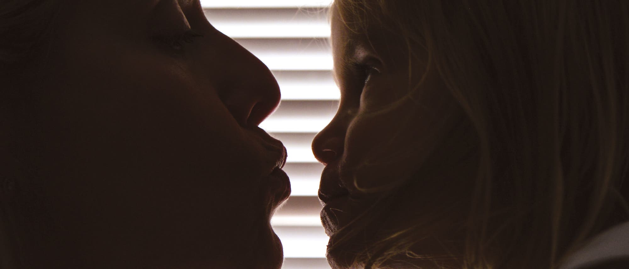 Eine Frau und ihr kleines Kind sehen sich an; die Gesichter sind im Profil zu sehen. Im Hintergrund sind horizontale Jalousien, die das Licht filtern und eine Silhouette erzeugen. Die Szene vermittelt Zuneigung und N&auml;he.