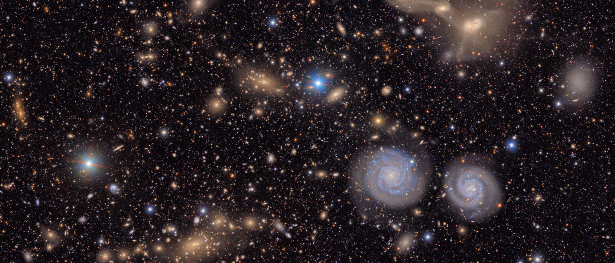 Eine dichte Ansammlung von Galaxien im Weltraum, darunter zwei markante Spiralgalaxien in der unteren rechten Bildhälfte. Der Hintergrund ist mit zahlreichen Sternen und weiteren, teils verschwommenen Galaxien gefüllt. Die Szene zeigt die Vielfalt und Weite des Universums.
