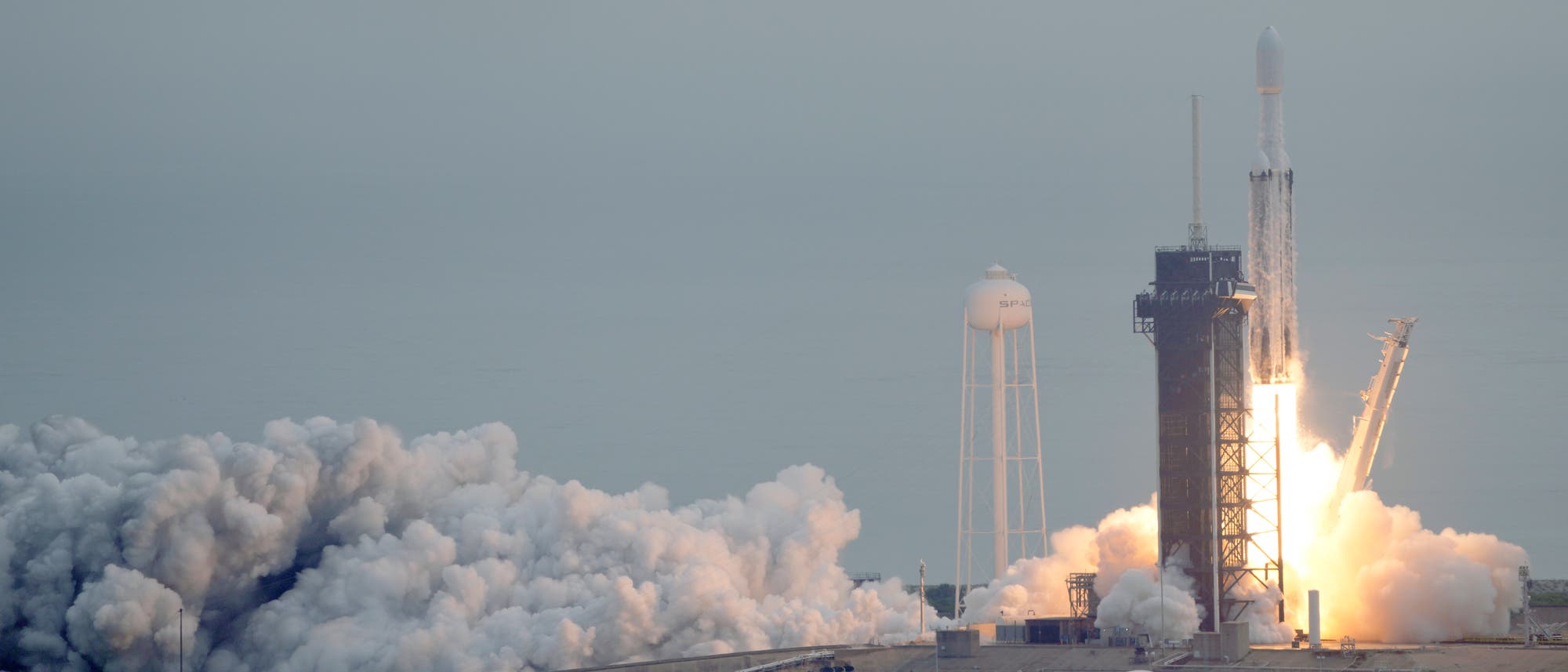 Die Rakete Falcon Heavy hebt vom Raumfahrtbahnhof  Kennedy Space Center in Florida ab. Der Himmel ist grau, der Raketentreibstoff verbrennt orange, links im Bild sieht man Wolken des verbrannten Treibstoffs