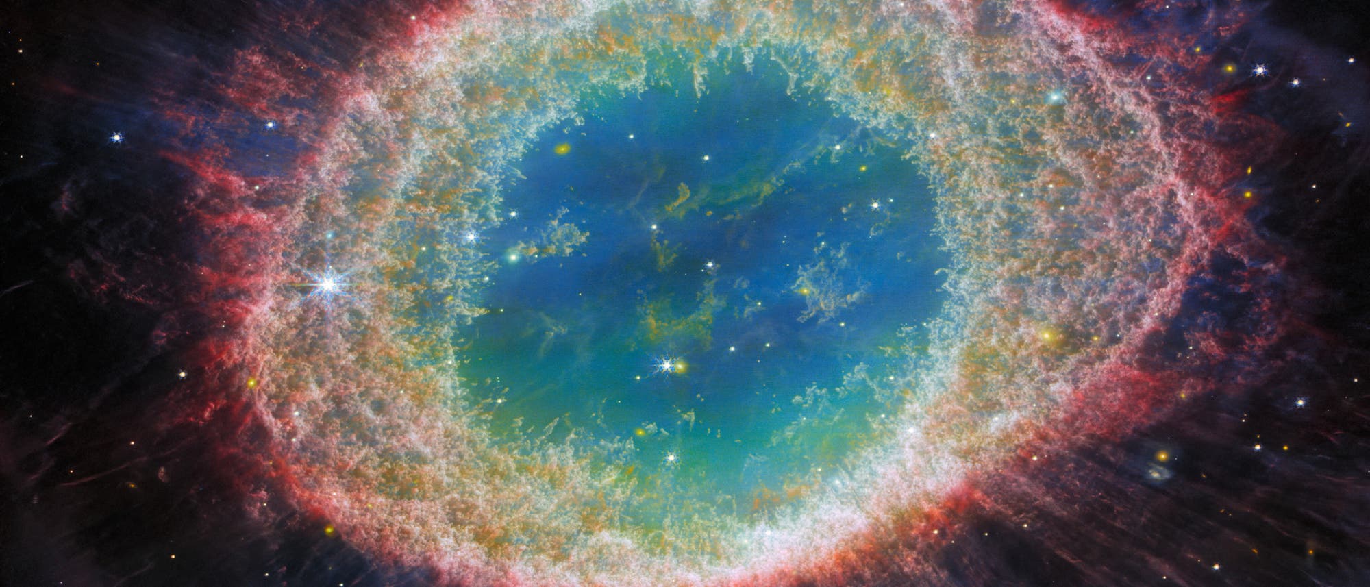 Ein farbenfrohes astronomisches Bild zeigt den Ringnebel im Weltraum. Im Zentrum befindet sich ein leuchtend blaues Gebiet, umgeben von einem mehrfarbigen, ringf&ouml;rmigen Nebel, der von Rot- und Gr&uuml;nt&ouml;nen dominiert wird. Der &auml;u&szlig;ere Bereich des Nebels zeigt feine, strahlenartige Strukturen, die in den dunklen Hintergrund des Universums &uuml;bergehen. Zahlreiche Sterne sind im Hintergrund sichtbar, was die Tiefe und Weite des Weltraums betont.