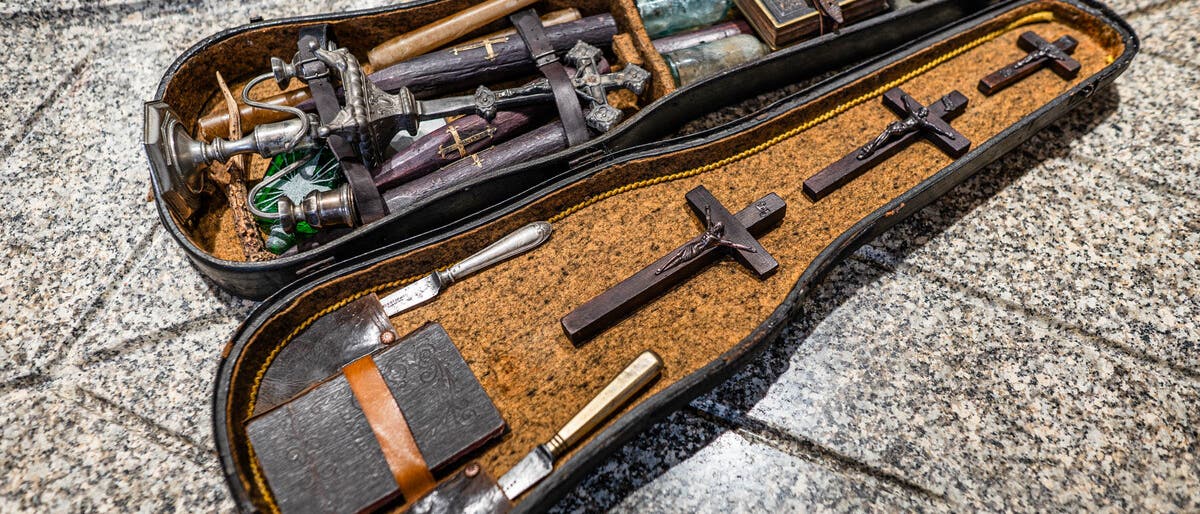 Ein offener Koffer auf einer Granitoberfl&auml;che zeigt eine Sammlung von Gegenst&auml;nden, darunter mehrere Kreuze, Flaschen, ein Buch und Messer. Die Gegenst&auml;nde sind sorgf&auml;ltig in einem mit Stoff ausgekleideten Innenraum angeordnet. Der Koffer scheint antik zu sein und die Anordnung der Objekte deutet auf ein historisches oder rituelles Set hin.