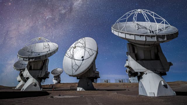 Eine Gruppe von großen Radioteleskopen steht auf einer trockenen, erdigen Fläche unter einem klaren Nachthimmel, der mit unzähligen Sternen übersät ist. Im Hintergrund ist die Milchstraße als leuchtendes Band sichtbar. Die Teleskopschüsseln sind in verschiedene Richtungen ausgerichtet und scheinen den Himmel zu beobachten.