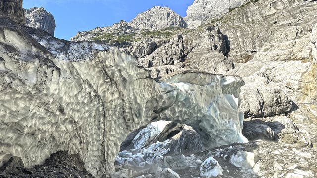 Eine natürliche Eishöhle in einer felsigen Berglandschaft. Die Höhle besteht aus blau-weißem Eis mit einer strukturierten Oberfläche. Im Hintergrund sind steile, graue Felswände und ein klarer blauer Himmel zu sehen. Die Szene vermittelt eine kühle, alpine Atmosphäre.