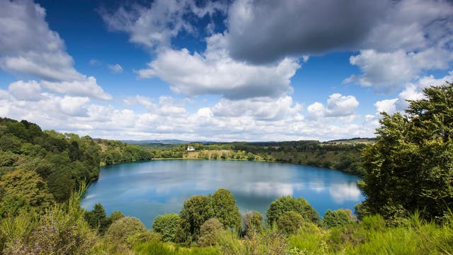 Ein malerischer See, umgeben von üppigen, grünen Bäumen und sanften Hügeln unter einem blauen Himmel mit weißen Wolken. Die Landschaft strahlt Ruhe und natürliche Schönheit aus. Im Hintergrund sind entfernte Hügel zu sehen.