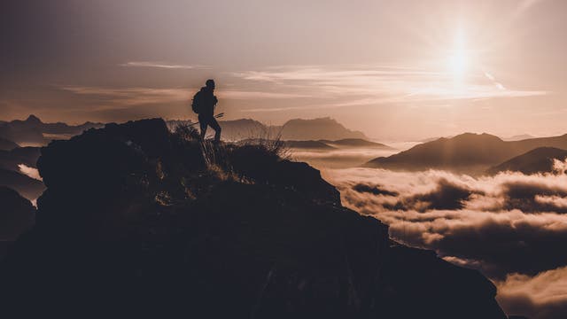 Eine Person steht auf einem felsigen Berggipfel und blickt in die Ferne. Der Himmel ist klar, und die Sonne geht über den Bergen auf, während Wolken die Täler darunter füllen. Die Szene vermittelt ein Gefühl von Abenteuer und Freiheit.