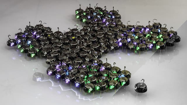 Eine Gruppe kleiner, runder Roboter mit leuchtenden LEDs in verschiedenen Farben (grün, blau, lila) formt eine dichte Formation auf einer glatten, reflektierenden Oberfläche. Ein einzelner Roboter ist etwas abseits von der Gruppe positioniert. Die Roboter sind gleichmäßig verteilt und scheinen miteinander zu interagieren, was auf eine koordinierte Bewegung oder ein gemeinsames Ziel hindeutet. Die Szene vermittelt ein Gefühl von Technologie und Zusammenarbeit.