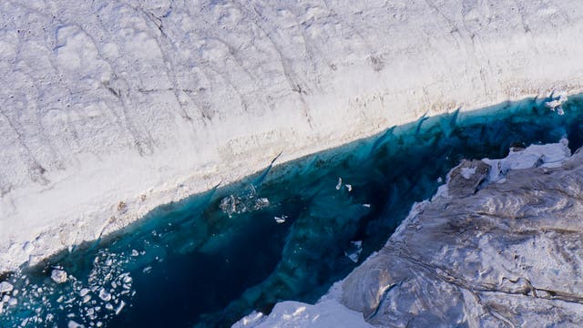 Luftaufnahme eines Gletschers mit schneebedeckter Oberfläche und einer tiefblauen Schmelzwasserlagune. Die weiße Schneedecke kontrastiert mit dem klaren, blauen Wasser, das von Eisschollen durchzogen ist. Die Struktur des Eises und die Farbverläufe im Wasser sind deutlich erkennbar.