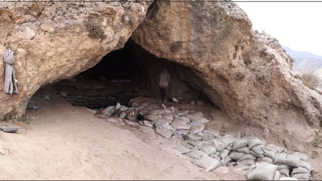 Eine große Höhle in einer felsigen Landschaft, deren Eingang mit Sandsäcken gesichert ist. Im Inneren der Höhle sind weitere Sandsäcke sichtbar. Eine Person steht in der Höhle, umgeben von Ausrüstungsgegenständen. Die Umgebung ist trocken und bergig, mit einem weiten Blick auf die umliegende Landschaft.