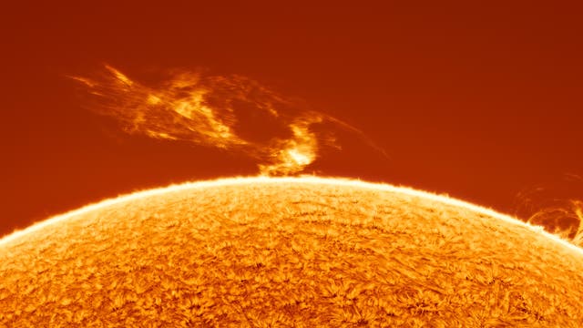 Eine Nahaufnahme der Sonnenoberfläche zeigt eine leuchtende, orangefarbene Textur mit zwei auffälligen Sonnenprotuberanzen, die sich in den Weltraum erstrecken. Die Protuberanzen erscheinen als helle, flammenartige Strukturen, die sich von der Sonnenoberfläche abheben. Der Hintergrund ist ein tiefes Orange, das die Intensität der Sonnenaktivität unterstreicht.