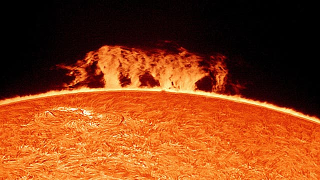 Die Abbildung zeigt eine Nahaufnahme der Sonnenoberfläche mit einer beeindruckenden Sonneneruption, die als leuchtende, flammenartige Struktur über dem Rand der Sonne sichtbar ist. Die Oberfläche der Sonne erscheint texturiert und zeigt die charakteristische Granulation. Der Hintergrund ist dunkel, was den Kontrast zur hellen Sonnenaktivität verstärkt. Schlüsselwörter: Sonne, Sonneneruption, Sonnenoberfläche, Astronomie.