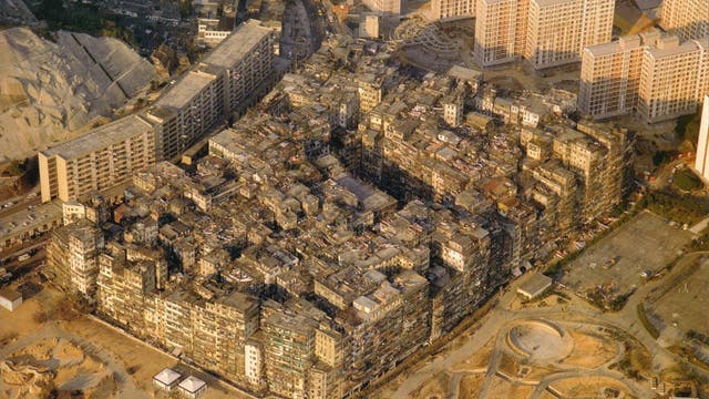 Luftaufnahme einer dichten städtischen Siedlung, umgeben von moderneren Hochhäusern und offenen Flächen. Die zentrale Struktur besteht aus eng aneinandergebauten, verwitterten Gebäuden, die ein labyrinthisches Muster bilden. Im Vordergrund sind geschwungene Wege und ein Parkplatz zu sehen. Die Szene zeigt den Kontrast zwischen alter und neuer Architektur.
