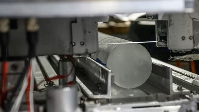 Eine Maschine schneidet einen zylindrischen Eisbohrkern. Der Eisbohrkern liegt in einer metallischen Halterung, während ein dünner Draht ihn durchtrennt. Die Umgebung wirkt industriell und technisch.