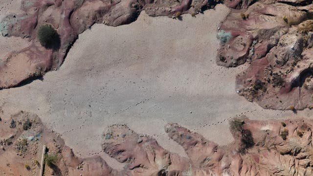 Luftaufnahme einer trockenen, felsigen Landschaft mit rötlichen und bräunlichen Tönen. In der unteren linken Ecke ist ein weißes Fahrzeug zu sehen. Die Oberfläche zeigt Spuren von Theropoden.