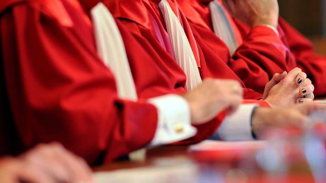 Eine Gruppe von Richterinnen und Richtern in roten Roben sitzt nebeneinander an einem Tisch. Die Gesichter sind nicht zu sehen, da das Bild nach oben abgeschnitten ist. Die Hände der Personen sind gefaltet oder ruhen auf dem Tisch. Die Roben haben weiße Kragen. Die Szene vermittelt einen formellen, gerichtlichen oder zeremoniellen Kontext.