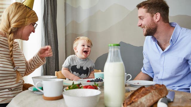 Mutter, Vater und Kleinkind lachen gemeinsam am Frühstückstisch