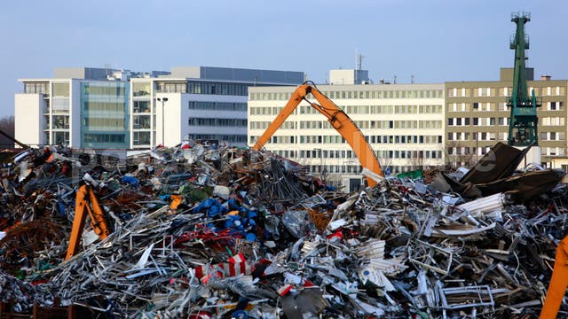 Ein großer Schrottplatz mit einem Haufen aus Metallabfällen, darunter bunte Teile und Schrottstücke. Zwei orangefarbene Kräne sind im Einsatz, um den Schrott zu bewegen. Im Hintergrund sind moderne Bürogebäude zu sehen, die den städtischen Charakter der Umgebung unterstreichen. Der Himmel ist klar und blau.
