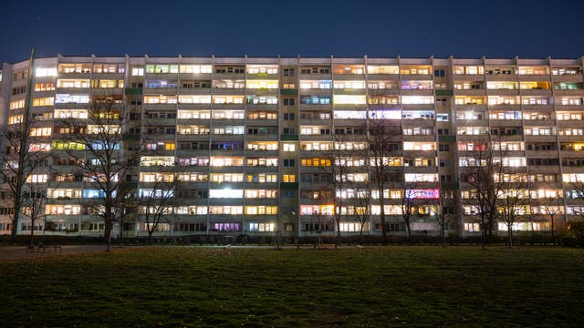 Ein mehrstöckiges Wohngebäude bei Nacht, dessen Fenster in verschiedenen Farben beleuchtet sind. Der Himmel ist klar und dunkel, und im Vordergrund ist eine Rasenfläche zu sehen. Einige Bäume stehen vor dem Gebäude. Die Szene vermittelt eine ruhige, nächtliche Atmosphäre.