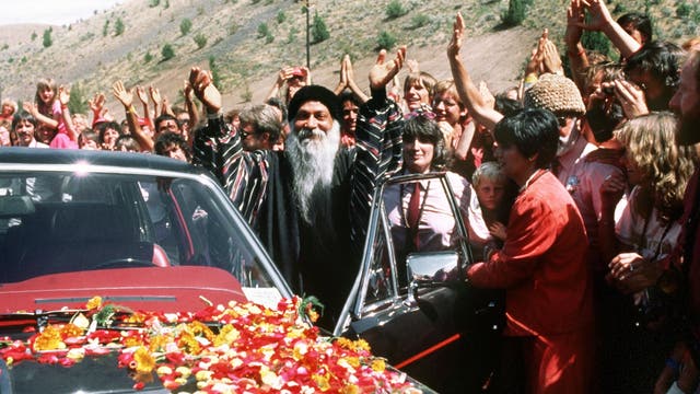 Eine Gruppe von Menschen steht um ein Auto, das mit Blumen bedeckt ist. Eine Person in der Mitte hebt die Arme, während die umstehenden Menschen klatschen und jubeln. Die Szene spielt sich in einer bergigen Landschaft ab. Die Stimmung wirkt feierlich und freudig.