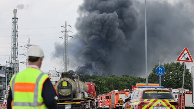 Einsatzfahrzeuge der Feuerwehr stehen am 27. Juli 2021 unweit einer Zufahrt zum Chempark, über dem eine dunkle Rauchwolke aufsteigt. 