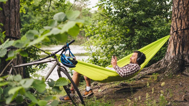 Eine Mann liegt entspannt in einer leuchtend grünen Hängematte zwischen zwei Bäumen und hört Musik über Kopfhörer. Er hält ein Smartphone in der Hand und lächelt. Neben der Hängematte steht ein Fahrrad mit einem blauen Helm am Lenker. Im Hintergrund ist ein Fluss und üppiges Grün zu sehen, was auf eine ruhige, natürliche Umgebung fern von Lärm und Arbeit hinweist.