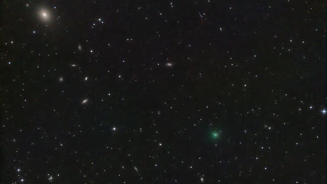 Ein astronomisches Bild zeigt einen sternenübersäten Nachthimmel mit mehreren Galaxien. Links oben ist eine helle, elliptische Galaxie zu sehen, umgeben von zahlreichen kleineren, unscharfen Galaxien. Über das Bild verteilt sind viele leuchtende Sterne unterschiedlicher Helligkeit. In der unteren rechten Bildhälfte ist ein schwach grünlich leuchtendes Objekt zu erkennen, möglicherweise ein Komet. Der Hintergrund ist tiefschwarz, was die Helligkeit der Himmelskörper hervorhebt.