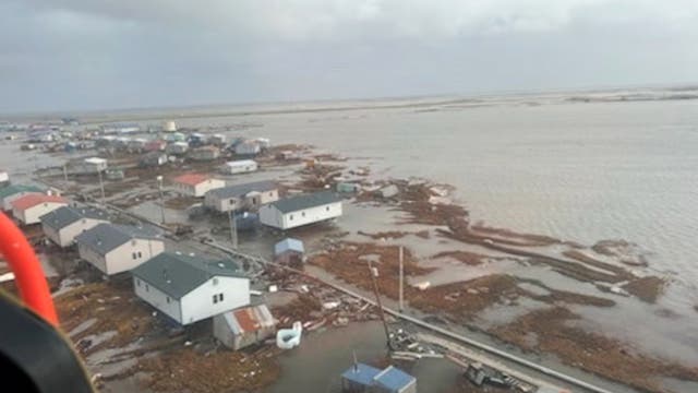 Luftaufnahme einer Küstenstadt, die von Überschwemmungen betroffen ist. Mehrere Häuser stehen in flachem Wasser, und die Straßen sind teilweise überflutet. Der Himmel ist bewölkt, was auf schlechtes Wetter hindeutet. Die Szene zeigt die Auswirkungen von Hochwasser auf die Infrastruktur der Stadt.