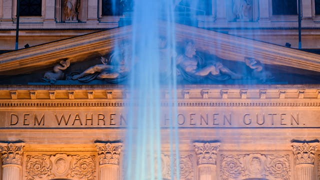 Das Bild zeigt das nächtlich beleuchtete Portal eines klassizistischen Gebäudes mit der großen Aufschrift »DEM WAHREN SCHOENEN GUTEN«. Im Vordergrund ist der Strahl eines Springbrunnens unscharf zu erkennen.