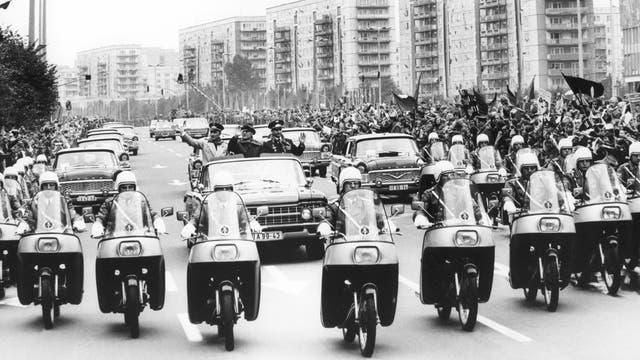 Angeführt von zahlreichen pfeilförmig formierten Polizeimotorrädern fahren mehrere Autos über die breite Karl-Marx-Allee, die von jubelnden Menschenmassen umsäumt ist. Im offenen Wagen an der Spitze sind winkend die Kosmonaten Sigmund Jähn und Waleri Bykowski neben DDR-Staatschef Erich Honecker zu sehen.