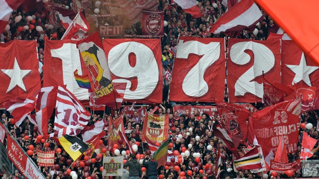 Eine große Menschenmenge in einem Fußballstadion hält rote Banner mit der weißen Aufschrift »1972« hoch. Um die Zahlen herum sind rote und weiße Fahnen und Schals zu sehen. Die Fans jubeln und schwenken rote und weiße Luftballons. Im Hintergrund sind weitere Fans und Fahnen zu erkennen.