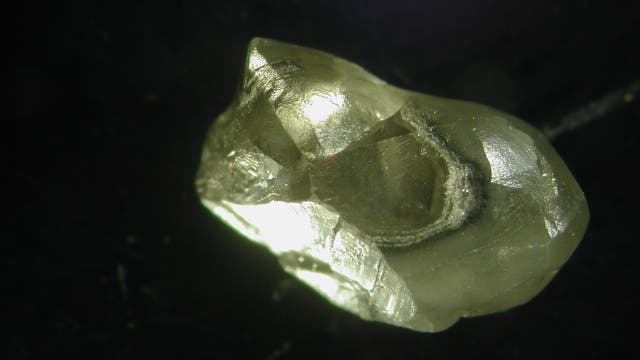 Nahaufnahme eines rohen, ungeschliffenen Diamanten auf schwarzem Hintergrund. Der Diamant zeigt eine unregelmäßige, kristalline Struktur mit glänzenden, reflektierenden Flächen. Die Lichtreflexionen betonen die Transparenz und die natürliche Form des Steins.