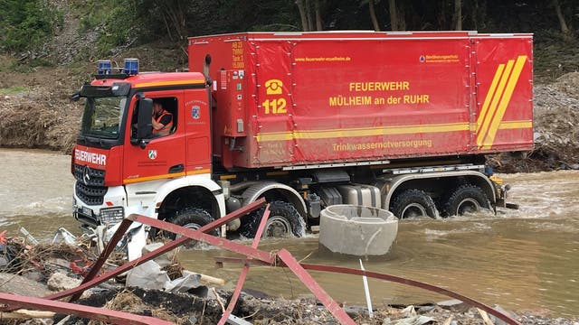 Ein Feuerwehrfahrzeug der Feuerwehr Mülheim an der Ruhr fährt durch ein überflutetes Gebiet. Auf dem roten Fahrzeug sind die Aufschrift "Trinkwasserversorgung" und die Notrufnummer 112 zu sehen. Im Vordergrund liegen Trümmerteile im Wasser. Im Hintergrund sind Bäume und ein schlammiger Hang zu erkennen.