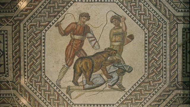Ein antikes Mosaik zeigt zwei Personen in römischer Kleidung, die mit Peitschen gegen einen Bären vorgehen. Der Bär steht über einer dritten Person, die am Boden liegt. Das Mosaik ist von einem komplexen geometrischen Muster umrahmt, das aus ineinander verschlungenen Bändern besteht. Die Szene vermittelt eine dynamische Darstellung von Bewegung und Spannung.