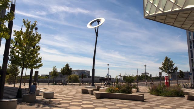 Vor einem fast klaren blauen Himmel auf einem öffentlichen Platz steht ein Kunstwerk, das aus einem 15 Meter hohen Metallpfahl besteht und einer an dessen Spitze aufgehängter, in der Sonne glänzender Metallscheibe, deren Durchmesser etwa vier Meter beträgt.