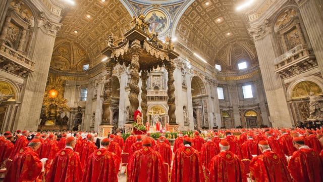 Eine große Versammlung von Kardinälen in roten Gewändern steht in der prunkvollen St. Petersbasilika im Vatikan. Der Innenraum ist reich verziert mit kunstvollen Säulen und einem beeindruckenden Baldachin über dem Altar. Die Kuppel ist mit kunstvollen Fresken geschmückt. Die Kardinäle sind nach vorne zum Altar ausgerichtet, wo eine religiöse Zeremonie stattfindet.