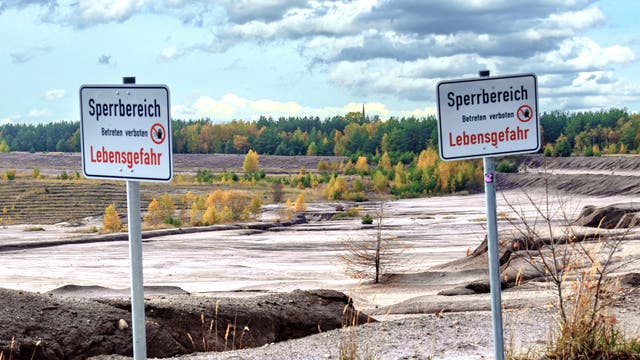 Warnschilder im Vordergrund, dahinter karge Sandlandschaft mit Erosionsrinnen, im Hintergrund ein Wald