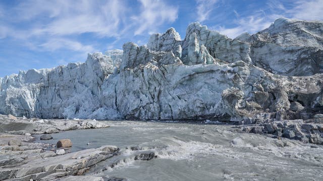 Gräuliches Schmelzwasser fließt über graues Gestein vor einer schmutzigweißen Eiswand in Grönland. Wenigstens ist der Himmel freundlich blau-weiß.