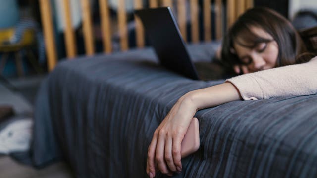 Eine schlafende, dunkelhaarige Frau liegt bekleidet auf dem Bett. Vor ihr steht ein aufgeklapptes Notebook, in der linken Hand, die aus dem Bett hängt, hält sie noch ihr Smartphone.
