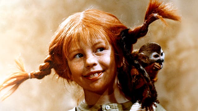 Ein Kind mit roten Zöpfen lächelt und schaut zur Seite. Auf der Schulter des Kindes sitzt ein kleiner Affe. Der Hintergrund ist unscharf und in warmen Tönen gehalten.