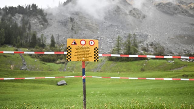 Ein gelbes Warnschild warnt davor, das dahinter liegende Gebiet zu betreten, weil ein Bergsturz droht. Verschwommen sieht man im Hintergrund den grauen Fels und eine Felsschutthalde. Beides ist umgeben von grünen Wiesen und Hängen.