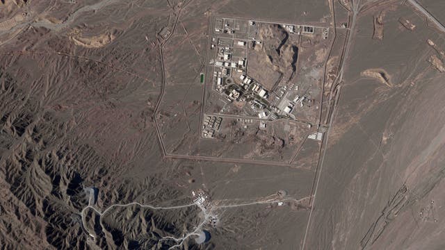 Satellitenbild einer großen, eingezäunten Anlage in einer Wüstenregion. Die Anlage besteht aus mehreren Gebäuden und Straßen, umgeben von kahlen, felsigen Hügeln. Es gibt keine sichtbaren Menschen oder Fahrzeuge. Die Umgebung ist trocken und karg, mit wenigen Vegetationsspuren.