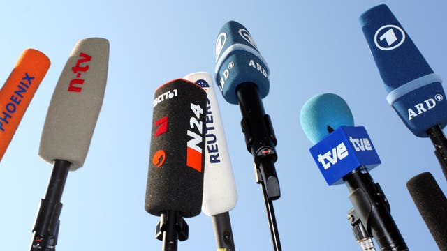 Mehrere Mikrofone verschiedener Nachrichtensender sind in die Luft gerichtet, darunter Phoenix, n-tv, N24, Reuters, ARD und tve. Die Mikrofone sind mit ihren jeweiligen Logos gekennzeichnet und stehen vor einem klaren blauen Himmel, was auf eine Pressekonferenz oder ein Medienereignis hindeutet.