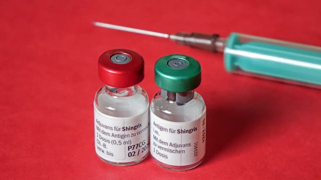 Zwei Impfstofffläschchen mit der Aufschrift „Adjuvans für Shingrix“ und „Antigen für Shingrix“ stehen nebeneinander auf einer roten Oberfläche. Die Fläschchen haben einen roten beziehungsweise grünen Verschluss. Im Hintergrund ist eine Spritze mit einer langen Nadel zu sehen.